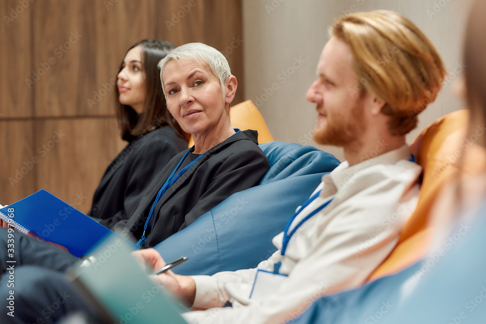 A lovable place for working. Middle-aged businesswoman looking at camera while making notes together with the audience at informal business meeting A lovable place for working. Middle-aged businesswoman looking at camera while making notes together with the audience at informal business meeting