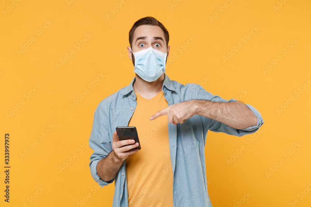 Shocked young man in sterile face mask posing isolated on yellow background in studio. Epidemic pandemic coronavirus 2019-ncov sars covid-19 flu virus concept. Pointing index finger on mobile phone.