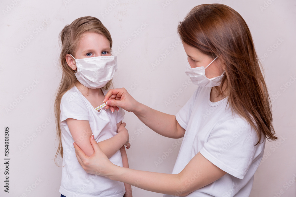 The concept of coronavirus quarantine. Young woman and her daughter in medical masks in isolation at home. Mom measures the temperature of her daughter. Theme of health and medicine