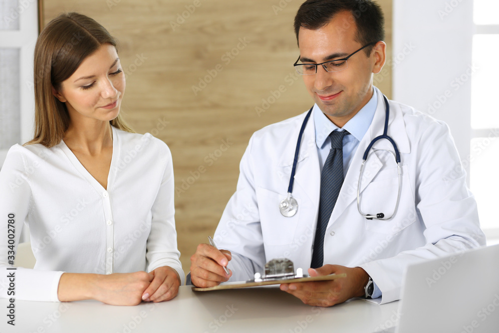 Doctor and patient discussing the results of a physical examination while sitting at a desk in a clinic. A male doctor using a clipboard to fill out a medical history of a young woman's medication