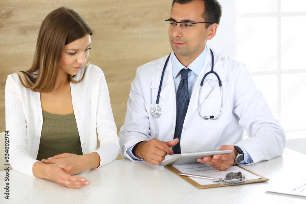 Doctor and patient discussing medical exam results while sitting at the desk in clinic. Male physician using tablet computer for filling up medication history records of young woman. Data in medicine Doctor and patient discussing medical exam results while sitting at the desk in clinic. Male physician using tablet computer for filling up medication history records of young woman. Data in medicine
