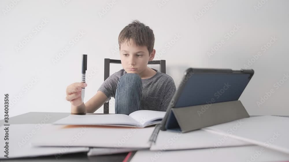 Sad child sitting at desk with tablet computer and many books. Boy feels bored, because he do not unterstand homework, he play with pen. Education, learning difficulties, online learning concept Sad child sitting at desk with tablet computer and many books. Boy feels bored, because he do not unterstand homework, he play with pen. Education, learning difficulties, online learning concept
