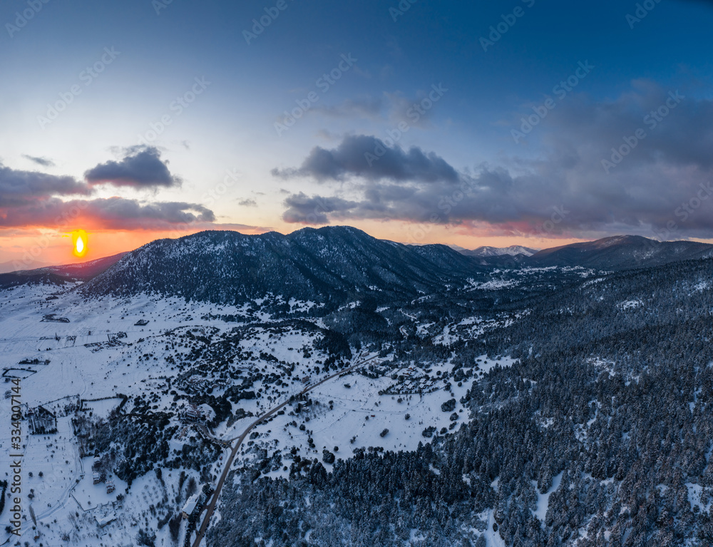 The Aerial view of a sunset over mountain in Arahova, Greece, a view of the valley below with trees covered by snow, colors of sunset The Aerial view of a sunset over mountain in Arahova, Greece, a view of the valley below with trees covered by snow, colors of sunset