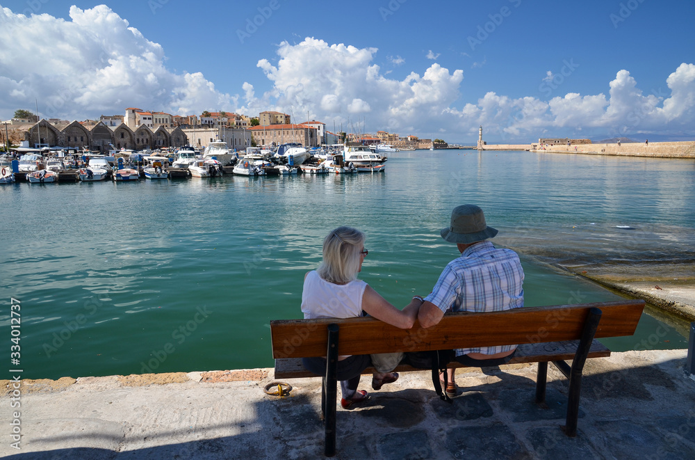 Unrecognizable elderly couple sitting on a bench and watching the wonderful view of Chania port in Crete island, Greece.