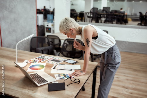 New project. Young stylish blonde tattooed female designer making some sketches and working with color swatch samples while standing near office desk photo