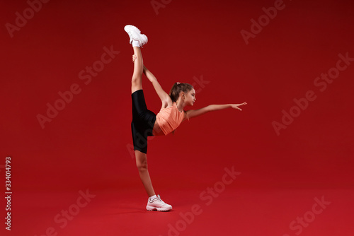 Strong beauty. A cute kid, girl is engaged in sport, gymnast is looking aside while stretching her legs. Isolated on red background. Fitness, training, active lifestyle concept. Horizontal shot © Svitlana