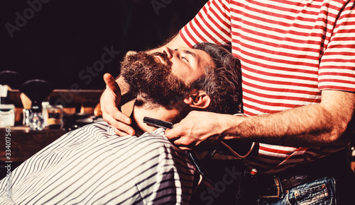 Barber shaving a bearded man in a barber shop. Bearded male in a barber shop while hairdresser shaves his beard with a dangerous razor. Straight razor, barbershop, beard. Vintage straight razor