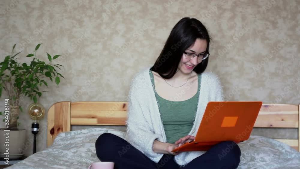 Young attractive girl works remotely while sitting on the bed at home. Young Cute girl works at a laptop while sitting on the bed. Work at home. Young attractive girl works remotely while sitting on the bed at home. Young Cute girl works at a laptop while sitting on the bed. Work at home.