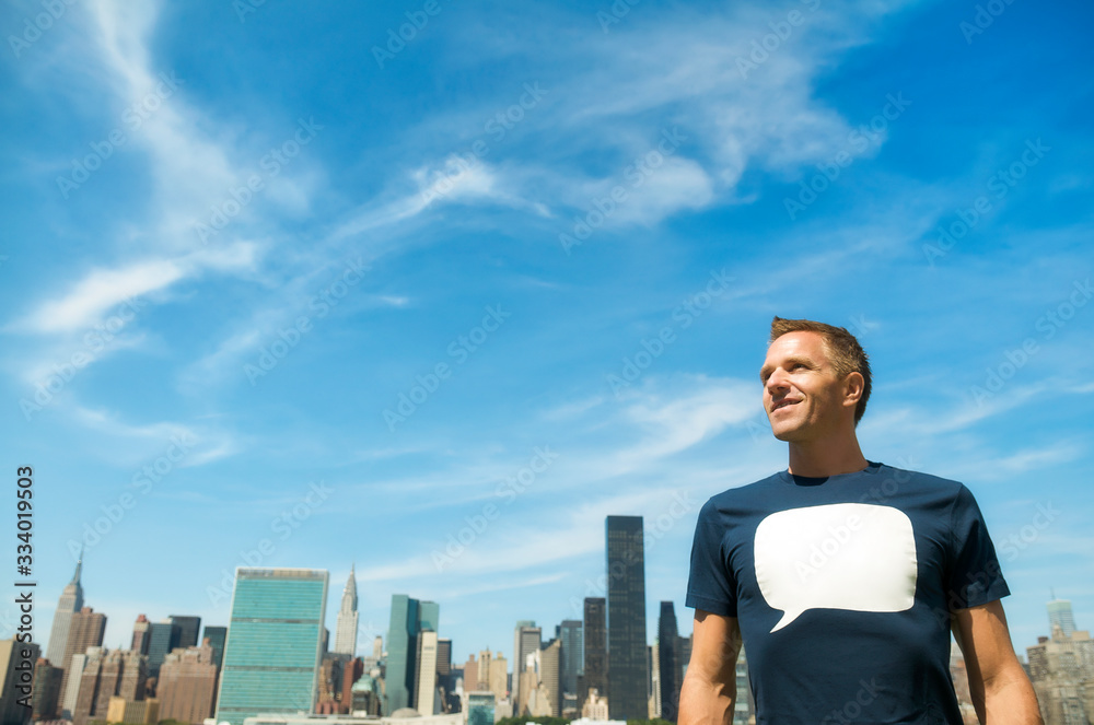 Smiling young man in a T-shirt with a blank speech bubble standing in front of the city skyline under bright blue sky