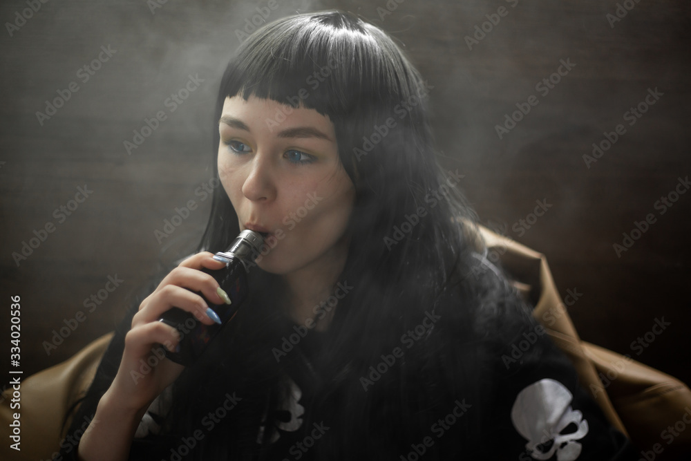 Fototapeta premium Vaping teenager. Young pretty white caucasian teenage brunette girl with blue eyes and problem skin smoking an electronic cigarette indoors. Deadly bad habit. Vape activity.