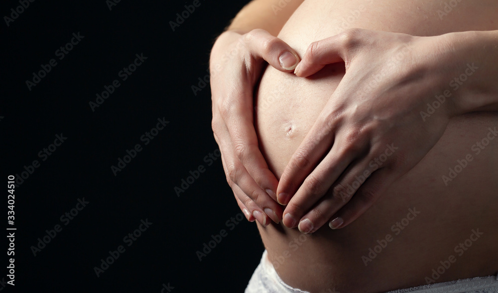 Pregnant Woman holding hands in a heart shape on her belly on black background