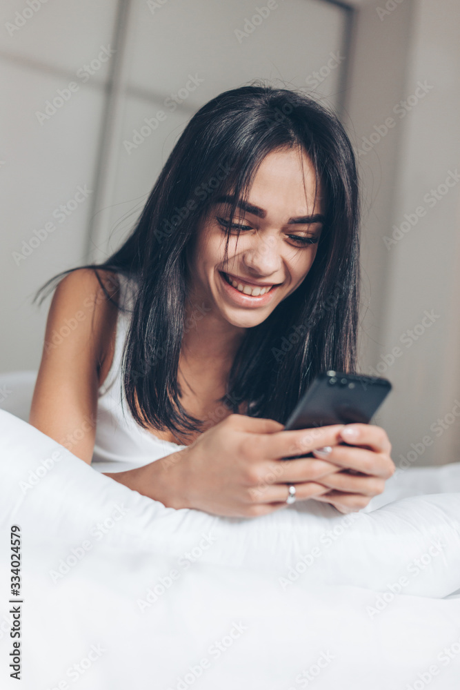 Happy Beautiful woman texting at smartphone while lying on her bed. Smiling Brunette Girl holding mobile phone Happy Beautiful woman texting at smartphone while lying on her bed. Smiling Brunette Girl holding mobile phone