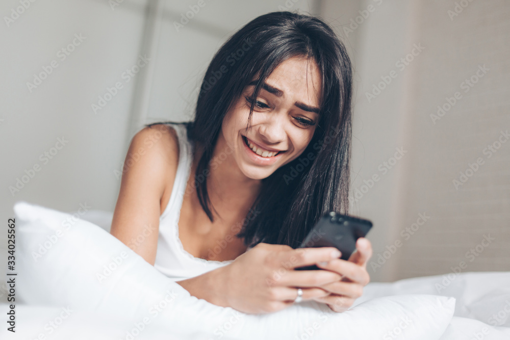 Happy Beautiful woman texting at smartphone while lying on her bed. Smiling Brunette Girl holding mobile phone Happy Beautiful woman texting at smartphone while lying on her bed. Smiling Brunette Girl holding mobile phone