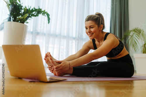 Close shot of blonde fit woman making forward fold yoga exercise while sitting on mat at home. Young woman is watching online yoga course due to COVID 19 pandemic.