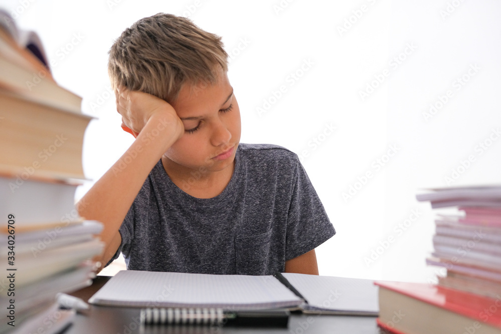 Schoolboy sitting among pile of books, textbooks, school exercise books on his desk and doing homework. Education, online distance learning, homeschooling during quarantine at home Schoolboy sitting among pile of books, textbooks, school exercise books on his desk and doing homework. Education, online distance learning, homeschooling during quarantine at home