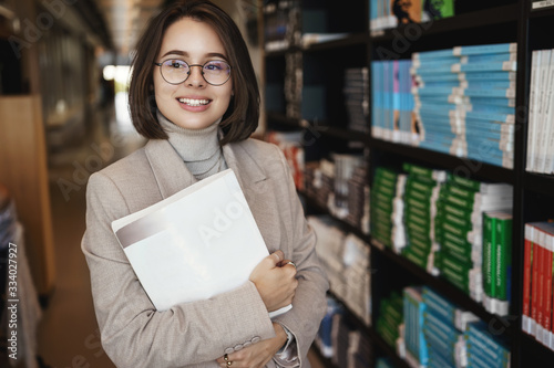 Portrait of young attractive, enthusiastic female tutor, teacher in elegant glasses and blazer, looking away with satisfied happy face, holding books, attend university library to pick material