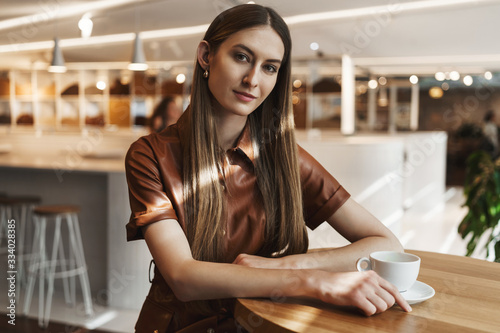 Empowered women concept. Attractive successful young businesswoman sitting in cafe, drinking cup coffee, tilt head and gazing camera with interest, smiling alluring