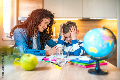 Mother Helping Son With Homework Sitting At Desk In kitchen. Mother helping son with homework. Boy having problems in finishing homework. Smiling mother helping adorable son doing schoolwork at home