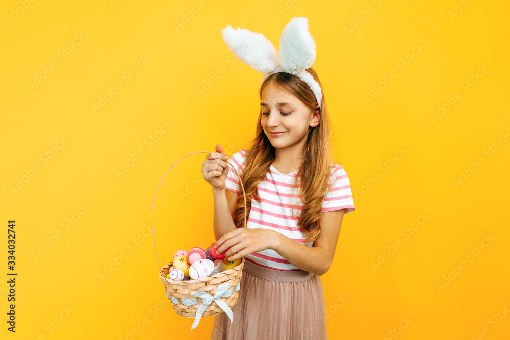 A beautiful happy girl on a head with rabbit ears, holding a wicker basket with colorful eggs on a yellow background