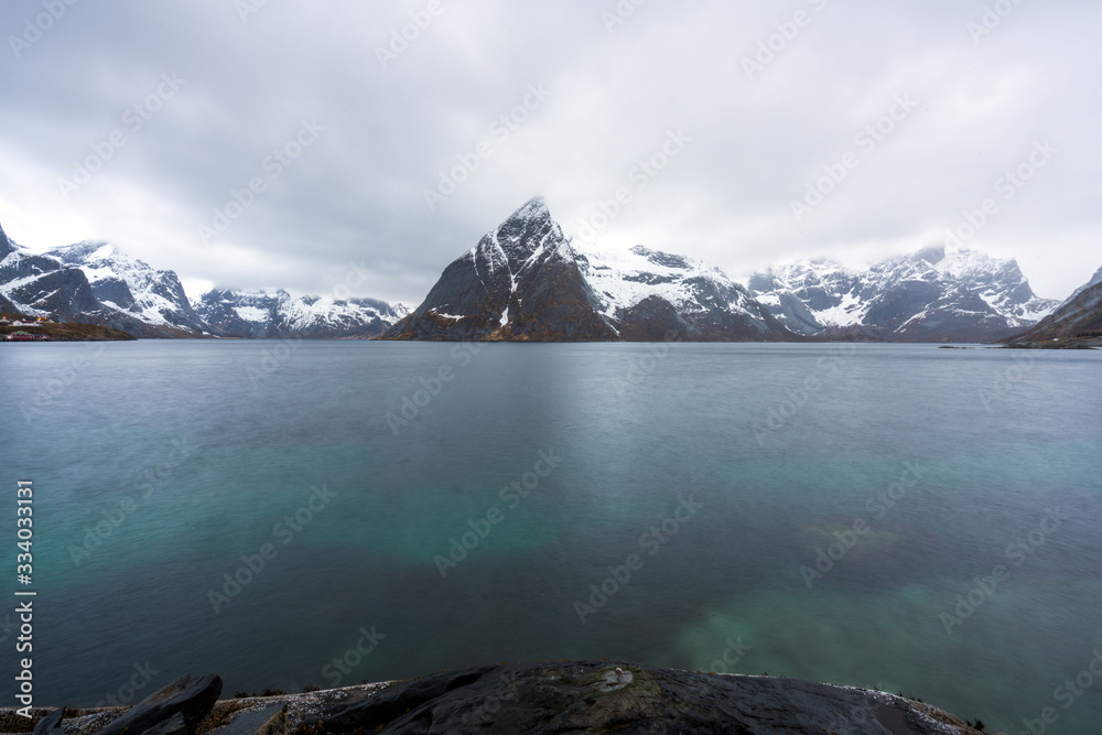 Mount Olstind peak in Lofoten, Norway with snow covered mountain chains. Long exposure shot to smooth out the green water infront. Traveling and destination concept.