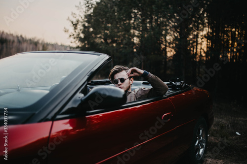 man in sunglasses sits in a red convertible in nature.
