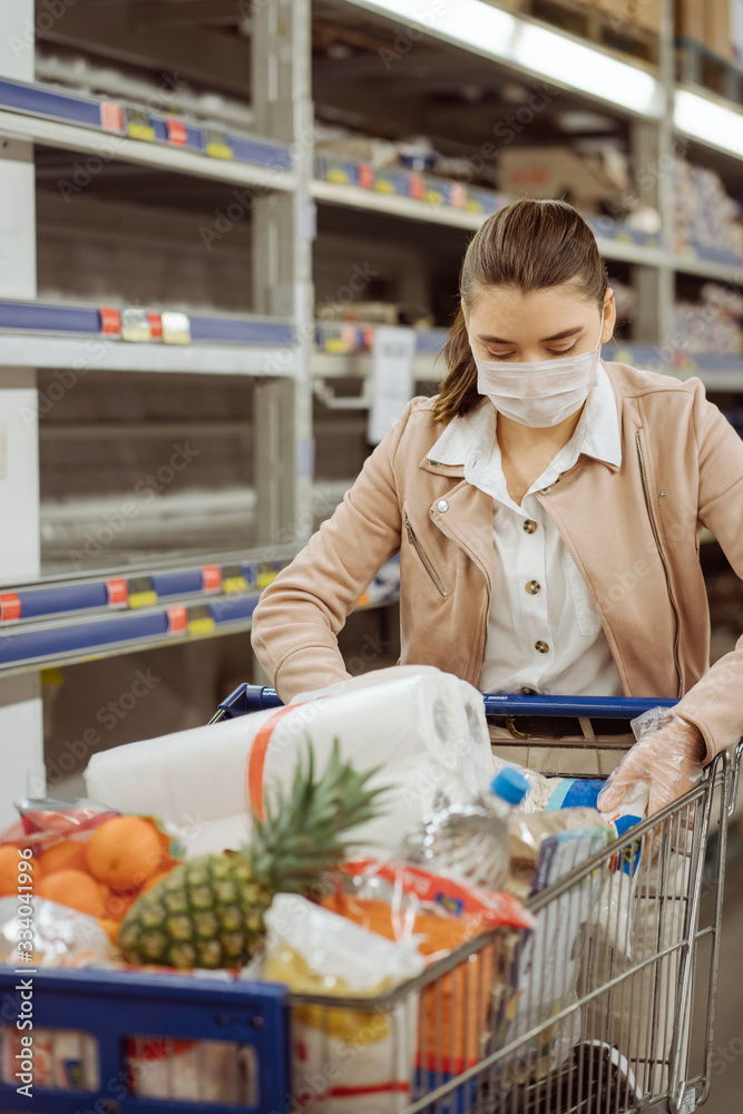 woman with medical facemask in supermarket puts groats in a basket.