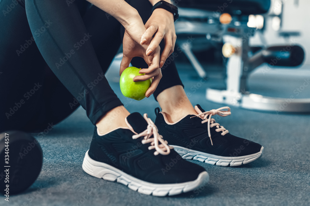 Naklejka premium Woman exercise workout in gym fitness break and relax. Hands holding apple fruit after training sport with dumbbell and water bottle beside her.