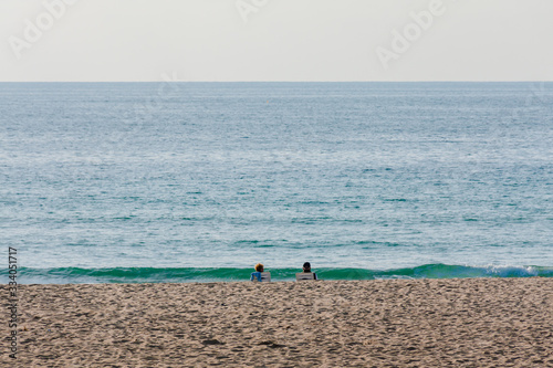 Two people sitting on a sandy beach in front of green wave coming out of blue ocean © BJ Clayden