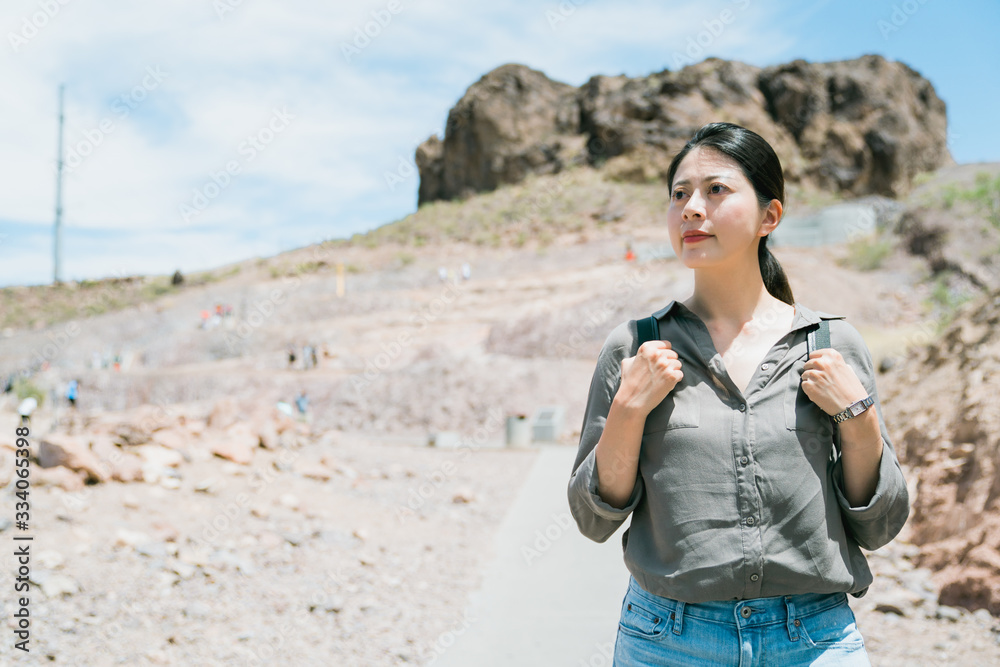 korean female backpacker gazing into distance background path up to a boulder. summer adventure to desert woman tourist hands on straps looking distant view on a dry land. korean female backpacker gazing into distance background path up to a boulder. summer adventure to desert woman tourist hands on straps looking distant view on a dry land.