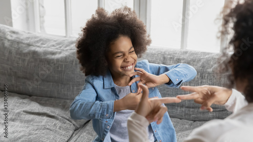 African deaf kid girl and her mother sitting on couch showing symbols with hands using visual-manual gestures enjoy communication at home. Hearing loss disability sign language learning school concept