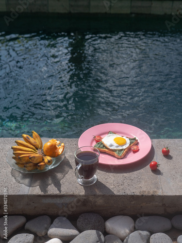 Breakfast, toasted bread with fried eggs, basil and cherry tomatoes on a pink plate, served by the pool.