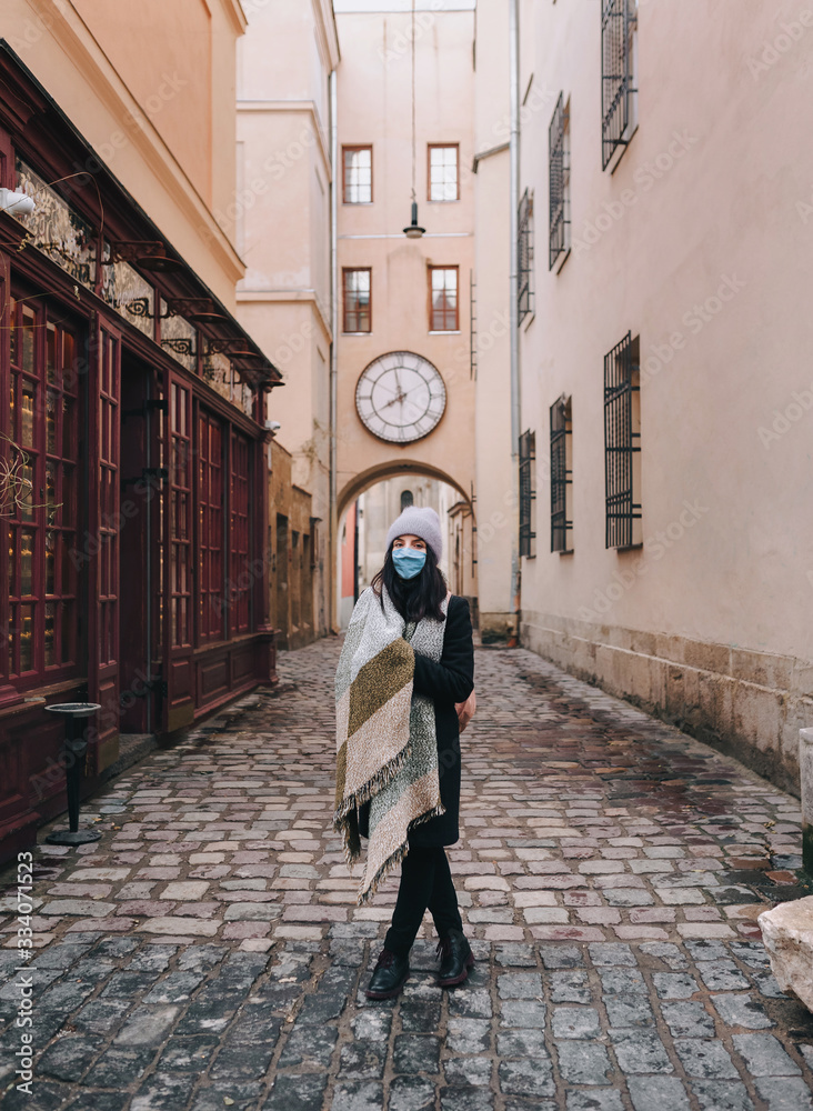 A young girl in a blouse, a scarf, boots and a protective medical blue mask is walking along the deserted narrow old street of the city. The concept of quarantine, coronavirus, pandemic COVID-19.