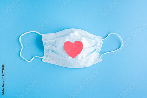 Disposable medical mask on blue background with red heart on it against bacteria and viruses. Stop the spread of coronavirus, self-defense. Copy space