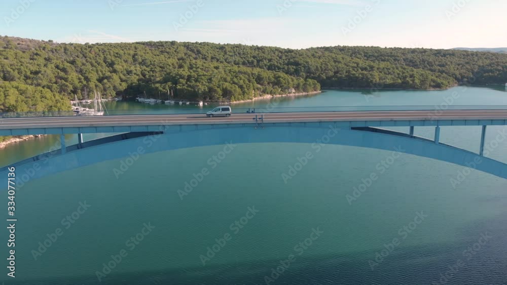 Aerial view lonely car driving fast on a concrete bridge over the ocean in a holiday paradise with wild nature and yacht parking on the beach