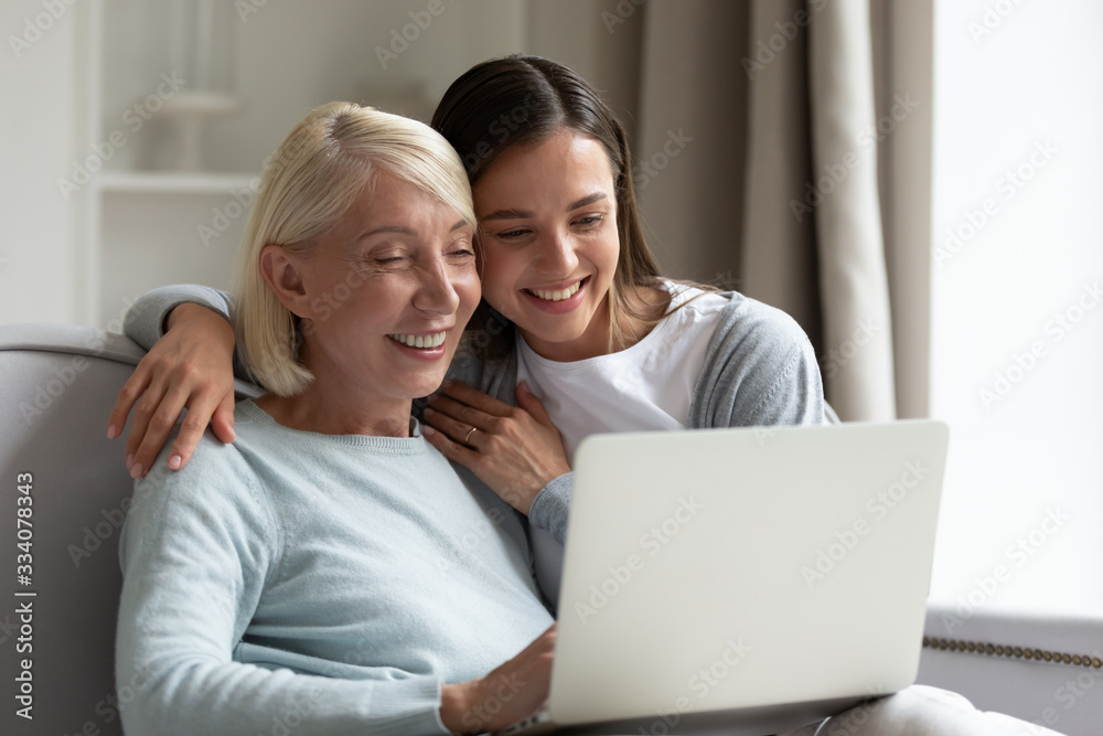 Smiling elderly mother and adult daughter sit relax on sofa in living room watch video on laptop together, happy senior mom and grown-up millennial girl child rest at home using modern computer Smiling elderly mother and adult daughter sit relax on sofa in living room watch video on laptop together, happy senior mom and grown-up millennial girl child rest at home using modern computer
