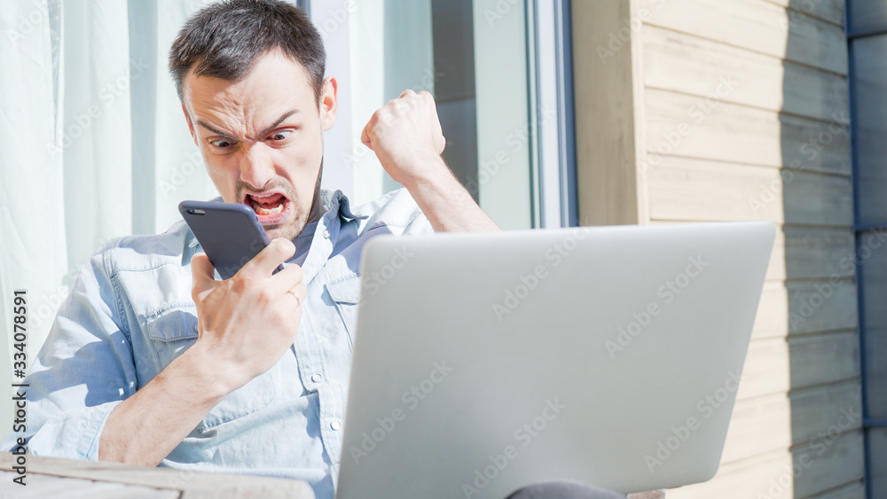 Angry businessman screaming on a phone while working behind computer from home. Stressful work environment concept. 
