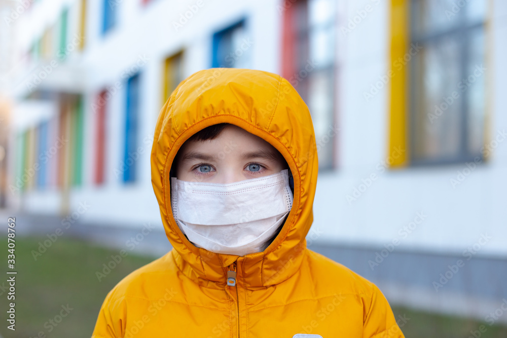 Portrait of a boy in a yellow jacket and hood in a white medical mask on the street