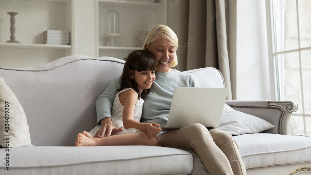 Happy elderly grandmother relax on couch in living room with little granddaughter watch video on laptop together, loving mature grandparent rest on sofa at home with small grandchild using computer Happy elderly grandmother relax on couch in living room with little granddaughter watch video on laptop together, loving mature grandparent rest on sofa at home with small grandchild using computer