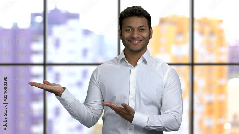 Young happy man shows empty copyspace with his both palms up. Wearing white formal shirt. Window background. Young happy man shows empty copyspace with his both palms up. Wearing white formal shirt. Window background.