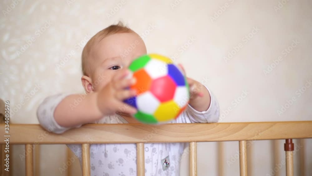 Cute Baby Playing With A Ball. Nine month old baby boy in a crib holds a colored ball. Child bites and throws a toy