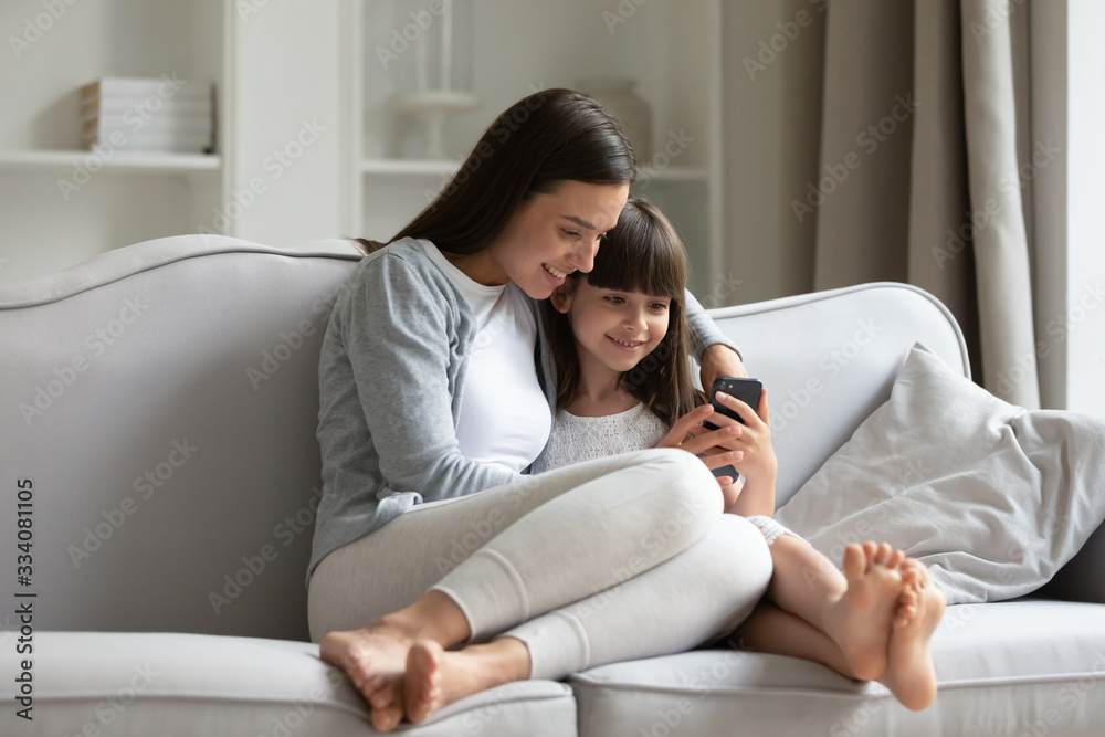 Happy young mother and cute little daughter relax on couch in living room using smartphone together, smiling millennial mom or nanny rest on sofa at home play on cellphone with small girl child Happy young mother and cute little daughter relax on couch in living room using smartphone together, smiling millennial mom or nanny rest on sofa at home play on cellphone with small girl child