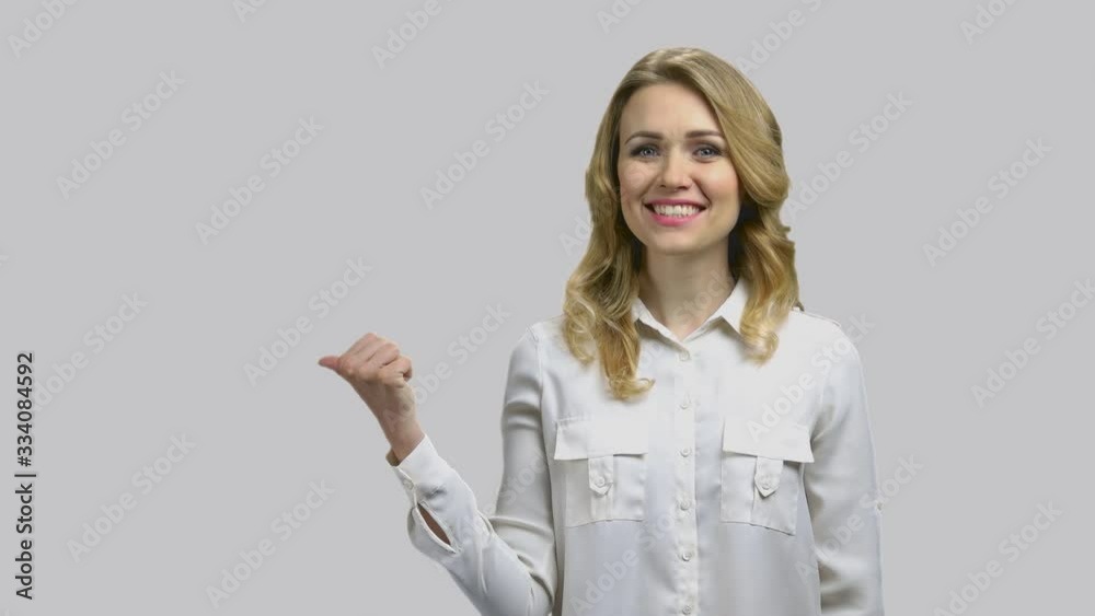 Young happy lady pointing back with her thumb. Blond cheerful woman attract your attention to something behind, grey background. Young happy lady pointing back with her thumb. Blond cheerful woman attract your attention to something behind, grey background.