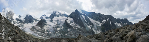 Panoramic snapshot of a ridge of snowy rocky mountains in Swiss Alps  sharp peaks are in stormy clouds  grey stones are on foreground