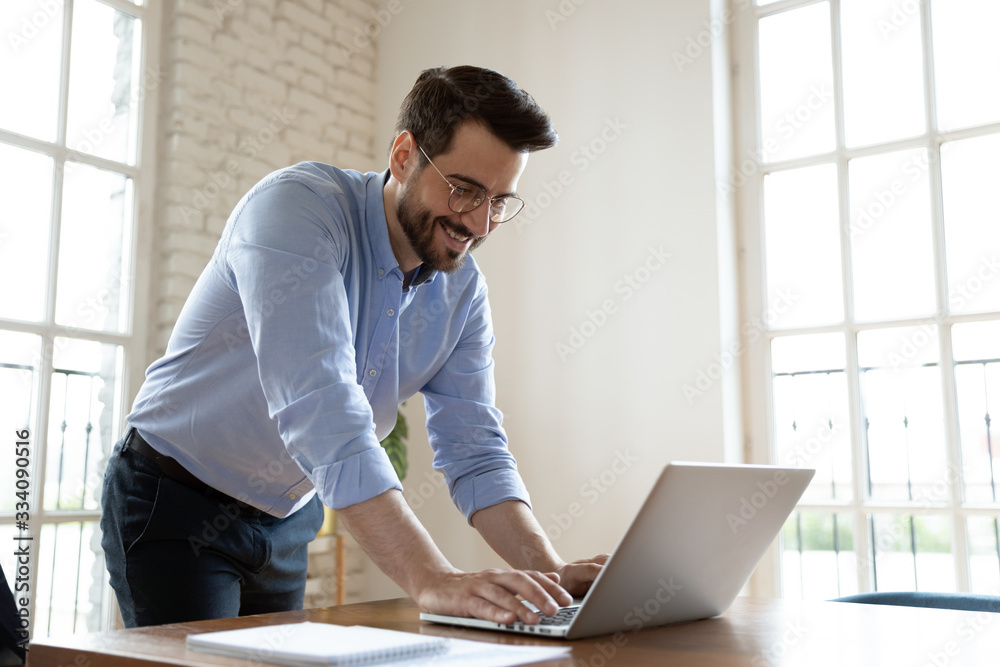 Millennial office employee leaning over table responds on business e-mail standing in boardroom, businessman ending workday chatting with friend on-line smiling having pleasant communication distantly