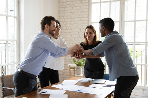 Four office workers mates gathered in boardroom putting stacked palms together showing support trust and unity taking part at team building activity having fun ending negotiations succeed common goal