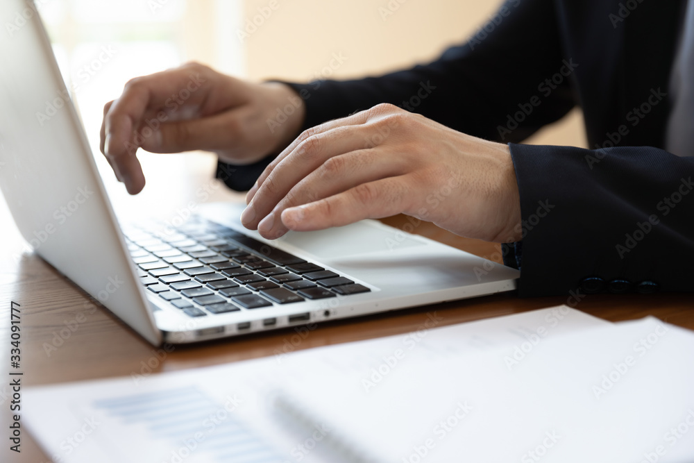 Businessman sit at desk typing on laptop, financial stats with charts lying on desk, hands device close up. Analyst working using pc develops insights, create business strategy. Office routine concept