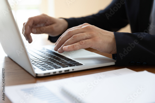 Businessman sit at desk typing on laptop, financial stats with charts lying on desk, hands device close up. Analyst working using pc develops insights, create business strategy. Office routine concept