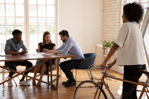 Diverse businesspeople talk sit at desk in office, focus on foreground African woman team leader came to meeting by bicycle, healthy benefits fit lifestyle environmental friendly vehicle usage concept