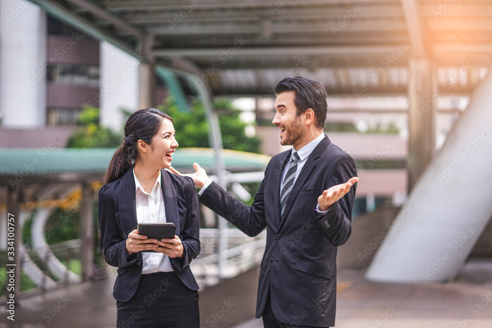 asian businesswoman holding a smart tablet device talking and laughing joyfully with a caucasian businessman, talking about business work or socializing, within urban city structure in the background asian businesswoman holding a smart tablet device talking and laughing joyfully with a caucasian businessman, talking about business work or socializing, within urban city structure in the background