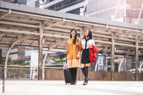 beautiful two asian women dressed casually holding each other arms smiling and laughing joyfully, holding a jacket in arm and dragging suitcase, walking through the urban city structure in background
